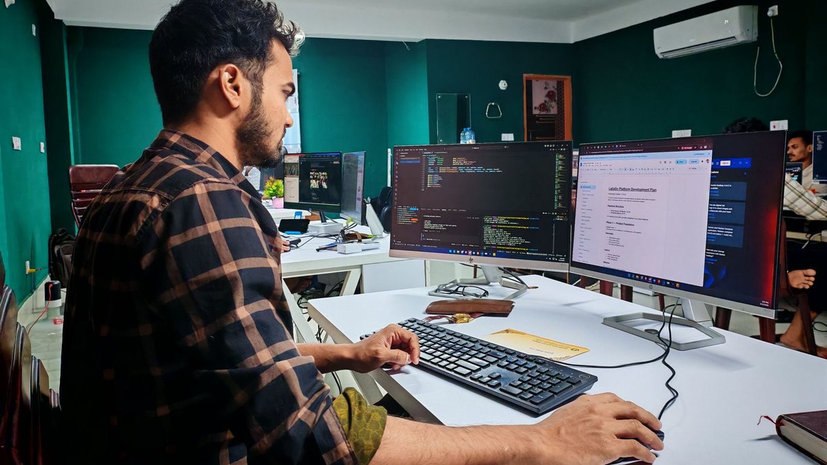 A developer at a desk in an office working across two large monitors, one showing code and one showing a project document