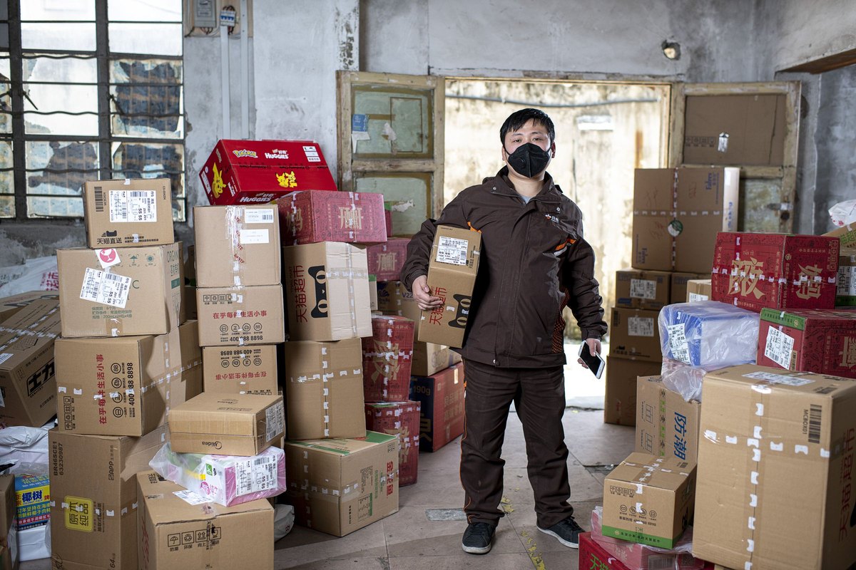 A masked courier stands among stacked parcels in a depot, holding a labelled package in one hand and a smartphone in the other