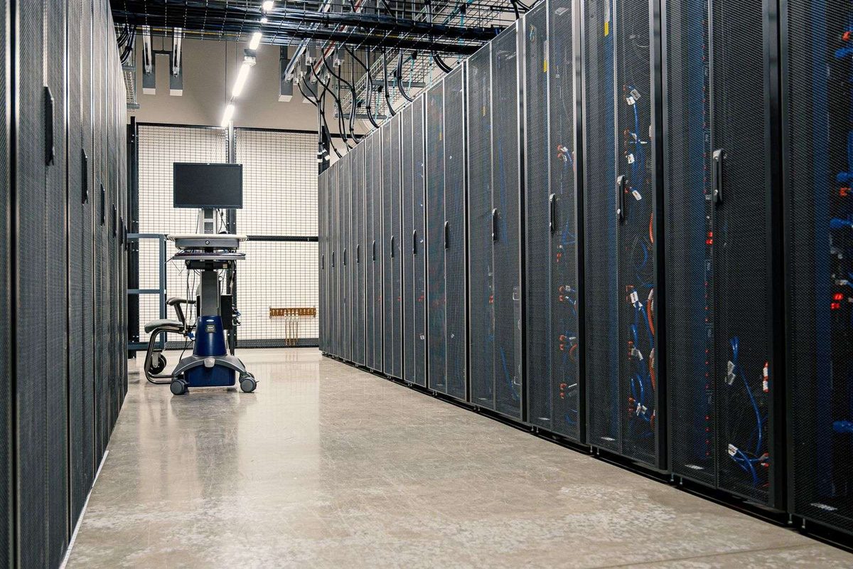 Data centre aisle with black mesh-door server racks, blue network cables and a monitor on a cart at the end of the row