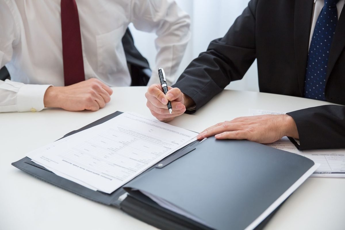 Two people in business suits at a desk, one signing a document in a leather folder
